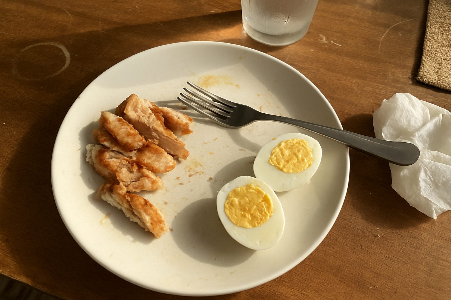 Porções de frango grelhado e ovos cozidos pela metade sobre prato já começado, com garfo apoiado sobre a comida e fundo de mesa de madeira arranhada